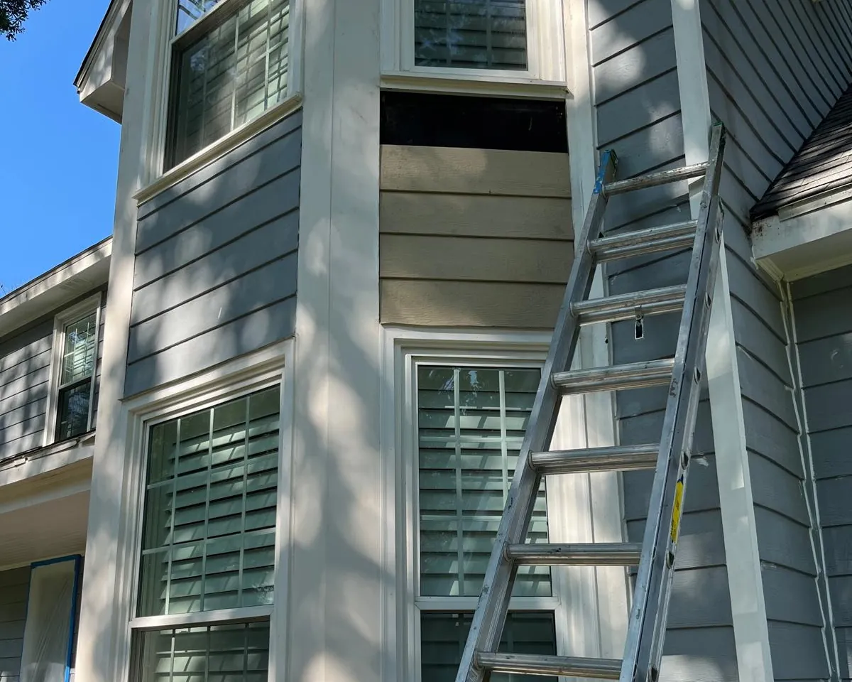 Tan lap siding set between two windows on a blue-grey house, with a dark gap left open above it and an aluminum ladder leaning on the wall